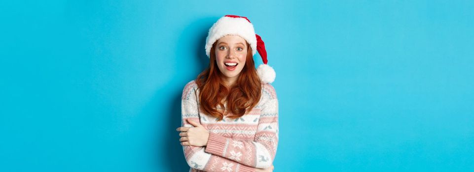 Winter and Christmas Eve concept. Amazed redhead girl in santa hat staring at camera in awe, smiling excited, standing over blue background.