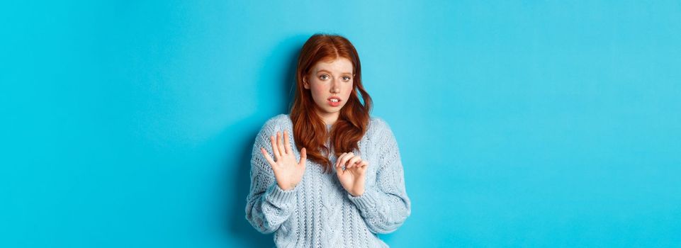 Worried redhead girl refusing or declining an offer, shaking hands and looking anxiously at camera, rejecting something unpleasant, standing over blue background.