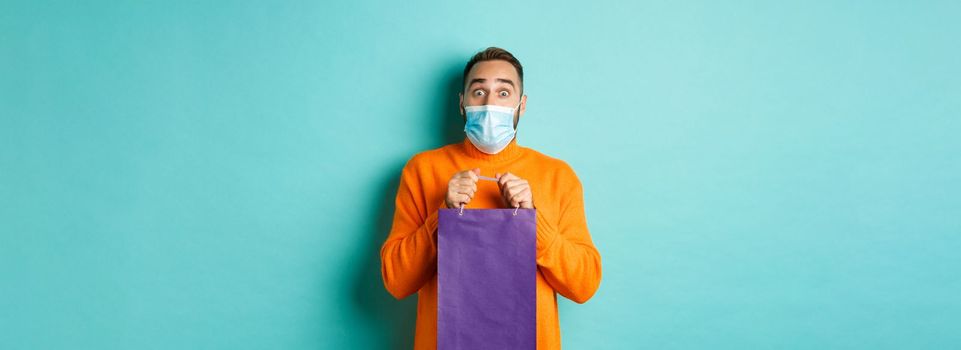 Covid-19, pandemic and lifestyle concept. Surprised man open shopping bag and looking amazed, receiving gift on holiday, standing over turquoise background.