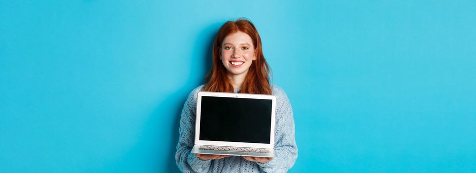 Young smiling woman with red hair and freckles showing computer screen, holding laptop and demonstrate online promo, standing over blue background.