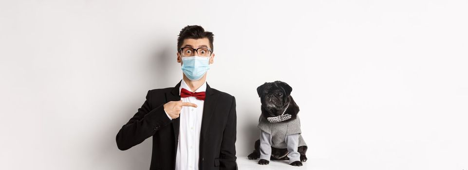 Coronavirus, pets and celebration concept. Amazed young man in face mask and suit pointing at cute black dog sitting near owner in party outfit, white background.