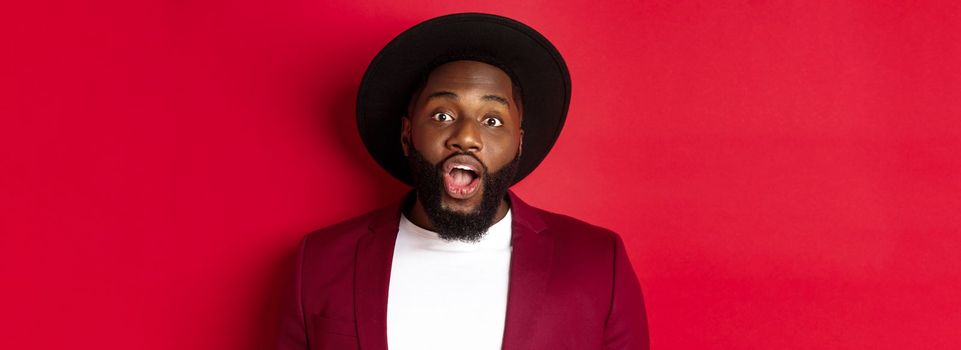 Close-up of impressed Black man with beard staring at camera, drop jaw and looking amazed, standing over red background.