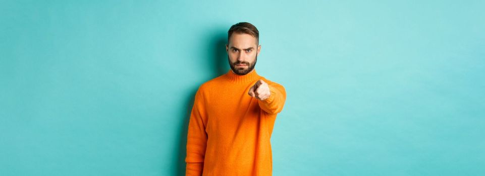 Confident man blame you, frowning and pointing finger at camera, accuse person, judgemental stare, standing over light blue background.