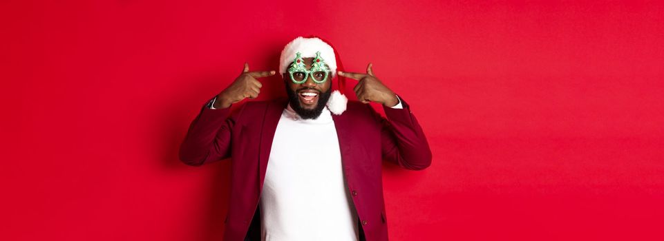 Merry Christmas. Cheerful Black man wearing funny party glasses and santa hat, smiling joyful, celebrating winter holidays, standing over red background.