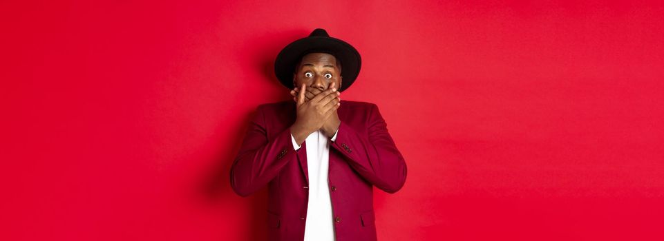 Christmas shopping and people concept. Shocked black man looking startled at camera, covering mouth with hands, standing against red background.