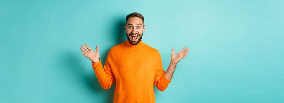 Excited man react to promo offer, spread hands sideways and rejoicing, standing against turquoise background in orange sweater.