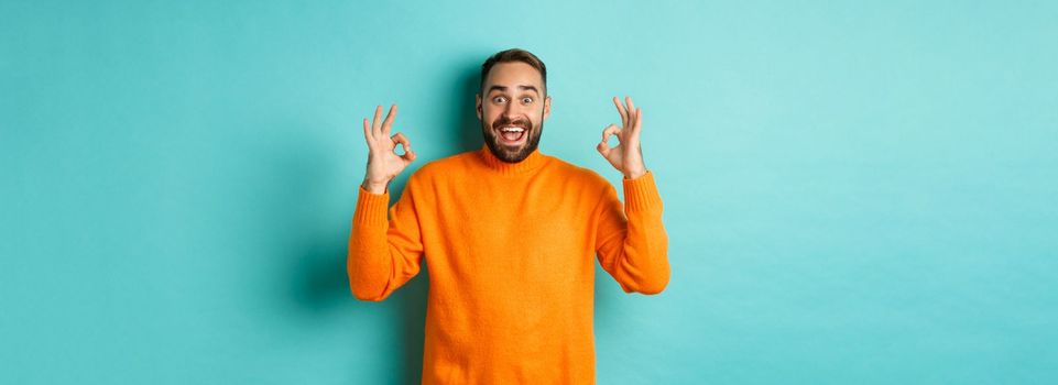 Impressed man praising something awesome, showing okay signs and looking amazed, standing in orange sweater at light blue background.