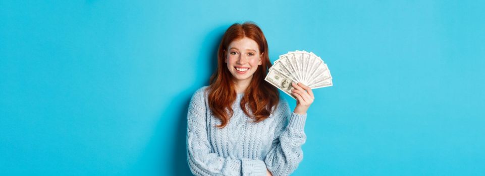 Happy and unbothered redhead woman holding money and smiling, looking confident and carefree, standing in sweater against blue background.