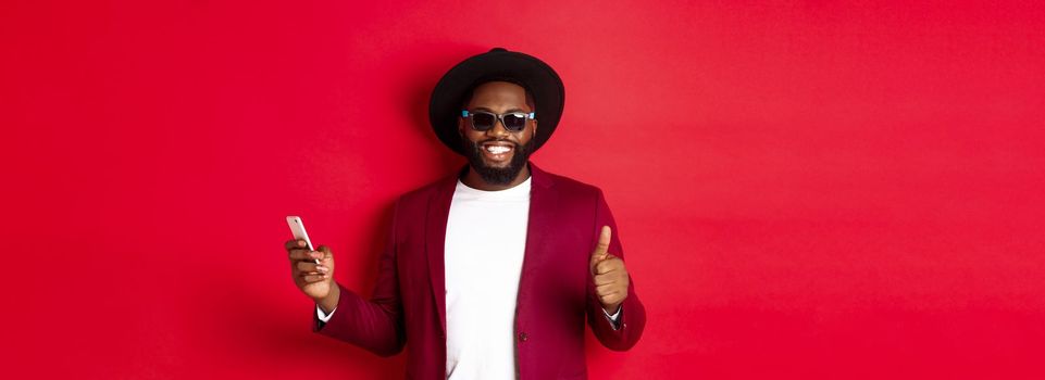 Handsome african american man using phone app and showing thumb up, smiling at camera, wearing sunglasses and fancy hat, red background.