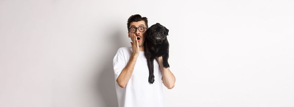 Image of shocked dog owner staring at camera and gasping impressed, cute black pug sitting on his shoulder and staring at you, white background.