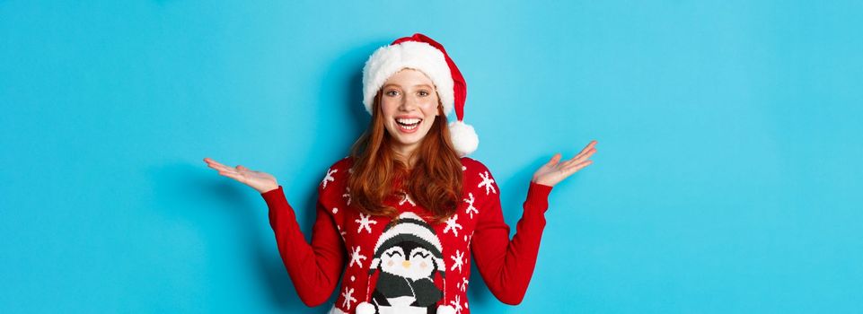 Happy holidays and Christmas concept. Cheerful redhead girl in Santa hat and xmas sweater, raising hands on copy spaces, holding something on blue background.