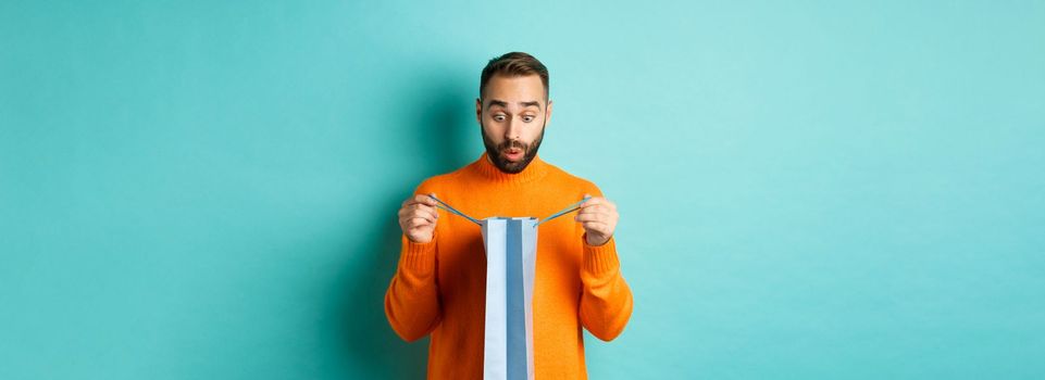 Surprised man open shopping bag and looking amazed, receiving gift on holiday, standing over turquoise background.