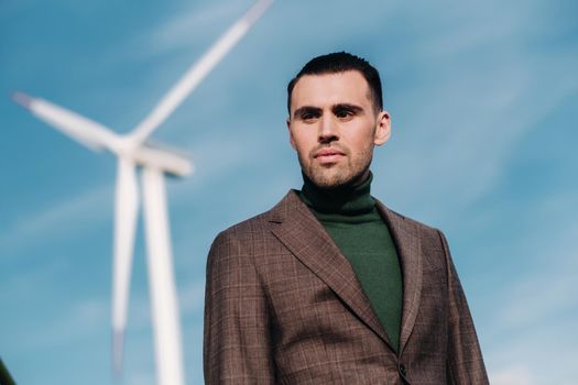 A man in a business suit with a green Golf shirt stands next to a windmill against the background of the field and the blue sky.Businessman near the windmills.Modern concept of the future