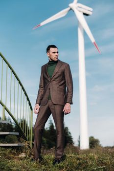 A man in a business suit with a green Golf shirt stands next to a windmill against the background of the field and the blue sky.Businessman near the windmills.Modern concept of the future