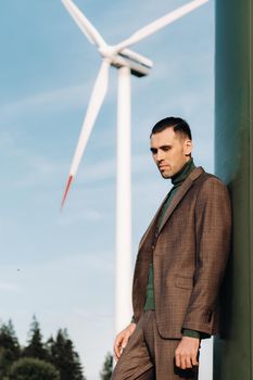 A man in a business suit with a green Golf shirt stands next to a windmill against the background of the field and the blue sky.Businessman near the windmills.Modern concept of the future