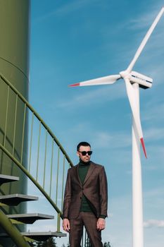 A man in a business suit with a green Golf shirt stands next to a windmill against the background of the field and the blue sky.Businessman near the windmills.Modern concept of the future