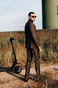 A man in a business suit and green Golf shirt stands next to an electric scooter against a field and blue sky.Businessman near the scooter.Modern concept of the future