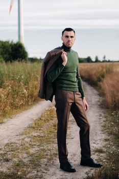 A man in a business suit with a green Golf shirt stands next to a windmill against the background of the field and the blue sky.Businessman near the windmills.Modern concept of the future