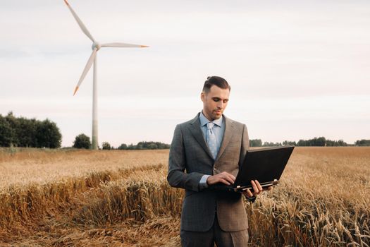 Portrait of a businessman in a suit holding a laptop in a field of wheat against the background of a windmill and the evening sky.