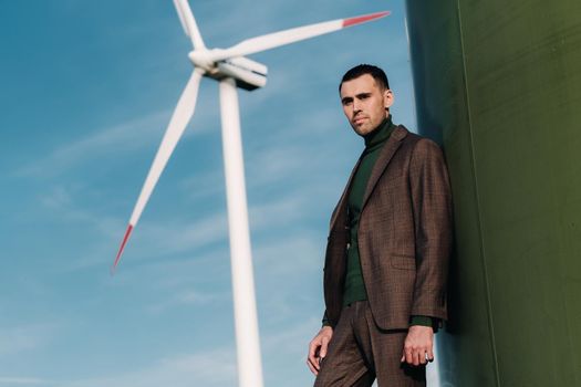 A man in a business suit with a green Golf shirt stands next to a windmill against the background of the field and the blue sky.Businessman near the windmills.Modern concept of the future