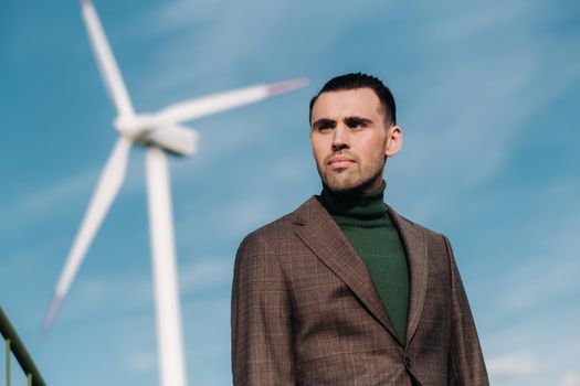 A man in a business suit with a green Golf shirt stands next to a windmill against the background of the field and the blue sky.Businessman near the windmills.Modern concept of the future