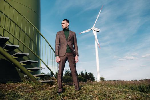 A man in a business suit with a green Golf shirt stands next to a windmill against the background of the field and the blue sky.Businessman near the windmills.Modern concept of the future