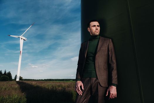 A man in a business suit with a green Golf shirt stands next to a windmill against the background of the field and the blue sky.Businessman near the windmills.Modern concept of the future