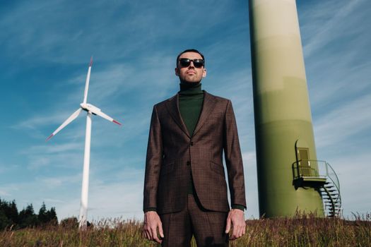 A man in a business suit with a green Golf shirt stands next to a windmill against the background of the field and the blue sky.Businessman near the windmills.Modern concept of the future