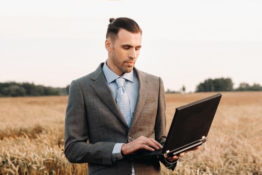 Portrait of a businessman in a suit holding a laptop in a field of wheat against the background of a windmill and the evening sky.