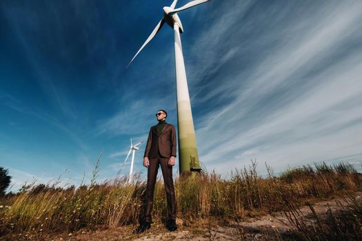 A man in a business suit with a green Golf shirt stands next to a windmill against the background of the field and the blue sky.Businessman near the windmills.Modern concept of the future