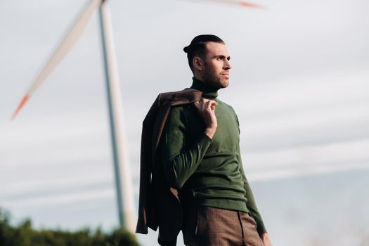 A man in a business suit with a green Golf shirt stands next to a windmill against the background of the field and the blue sky.Businessman near the windmills.Modern concept of the future