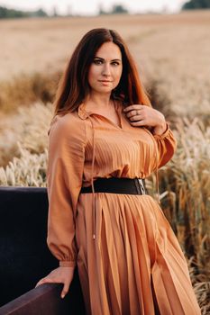 A girl in a orange dress stands next to a chair in a wheat field in the evening.