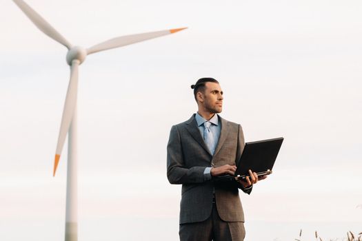 Portrait of a businessman in a suit holding a laptop in a field of wheat against the background of a windmill and the evening sky.