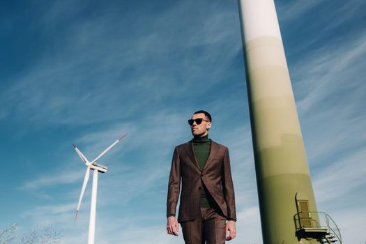 A man in a business suit with a green Golf shirt stands next to a windmill against the background of the field and the blue sky.Businessman near the windmills.Modern concept of the future