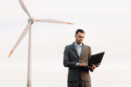 Portrait of a businessman in a suit holding a laptop in a field of wheat against the background of a windmill and the evening sky.