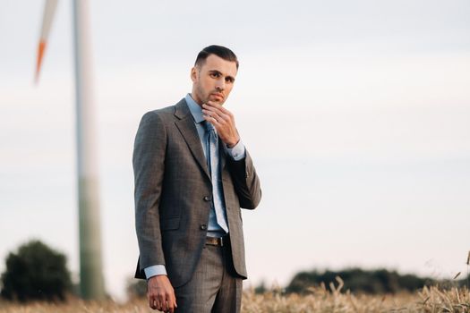 Portrait of a businessman in a gray suit on a wheat field against the background of a windmill and the evening sky.