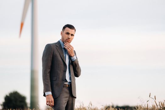 Portrait of a businessman in a gray suit on a wheat field against the background of a windmill and the evening sky.