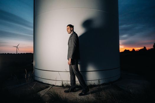 A man in a grey business suit stands next to a windmill after sunset .Businessman near windmills at night.Modern concept of the future