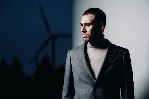 A man in a grey business suit stands next to a windmill after sunset .Businessman near windmills at night.Modern concept of the future