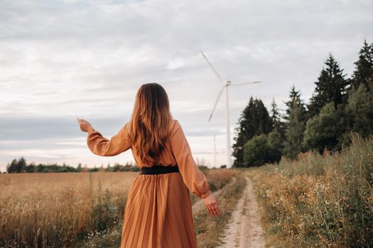 unrecognizable girl in a orange long dress with long hair in nature in the evening against the background of a forest and a windmill.