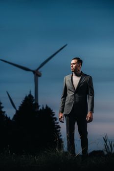 A man in a grey business suit stands next to a windmill after sunset .Businessman near windmills at night.Modern concept of the future