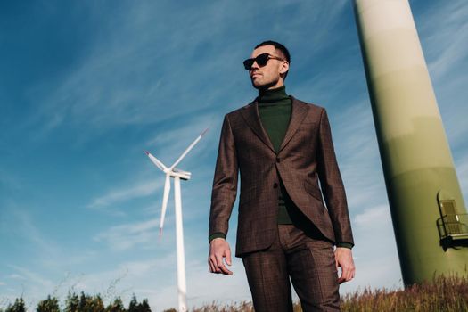 A man in a business suit with a green Golf shirt stands next to a windmill against the background of the field and the blue sky.Businessman near the windmills.Modern concept of the future