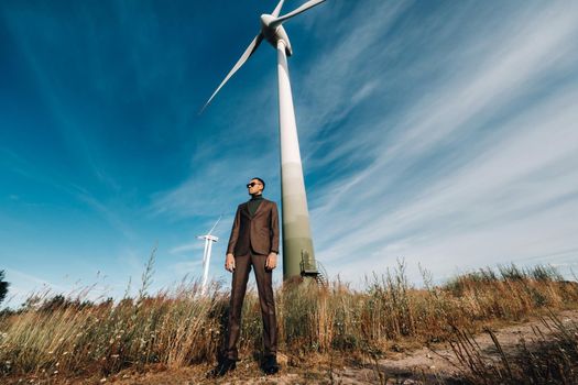 A man in a business suit with a green Golf shirt stands next to a windmill against the background of the field and the blue sky.Businessman near the windmills.Modern concept of the future