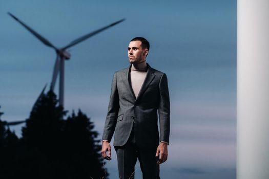 A man in a grey business suit stands next to a windmill after sunset .Businessman near windmills at night.Modern concept of the future