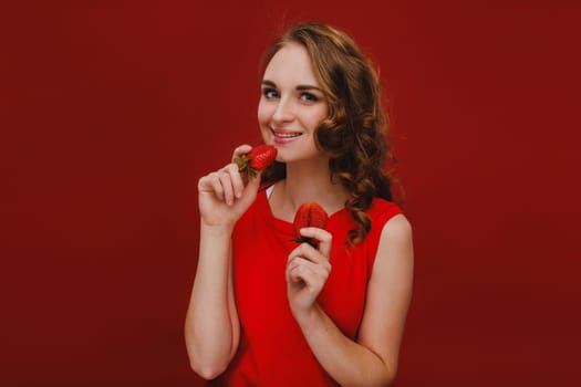 A beautiful girl in a red dress on a red background holds a strawberry in her hands and smiles.