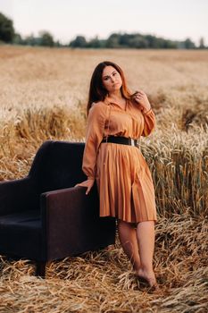 A girl in a orange dress stands next to a chair in a wheat field in the evening.
