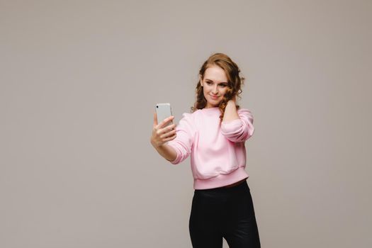 A smiling happy girl in a pink blouse takes a selfie on a smartphone on a gray background