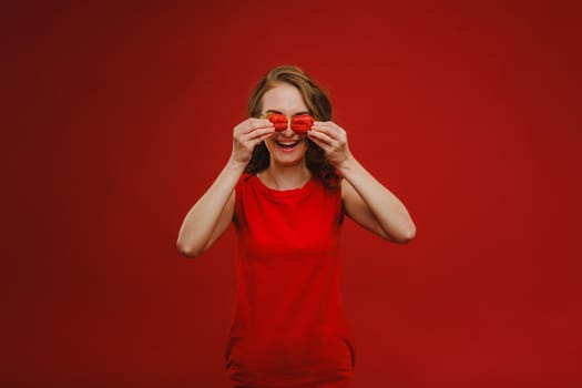 A beautiful girl in a red dress on a red background holds a strawberry in her hands and smiles.
