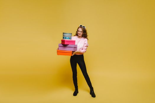 A happy young woman with colorful paper boxes after shopping isolated on an orange Studio background. Seasonal sales, purchases, spending money on gifts.