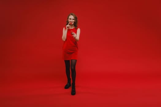 A beautiful girl in a red dress on a red background holds a strawberry in her hands and smiles.